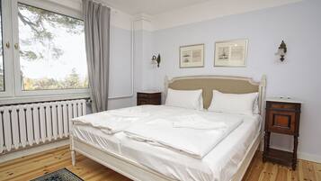 Ein ordentlich bezogenes Bett in einem Zimmer mit Heizkörper, einem fenster mit Vorhängen und zwei gerahmten Bildern an der Wand.
