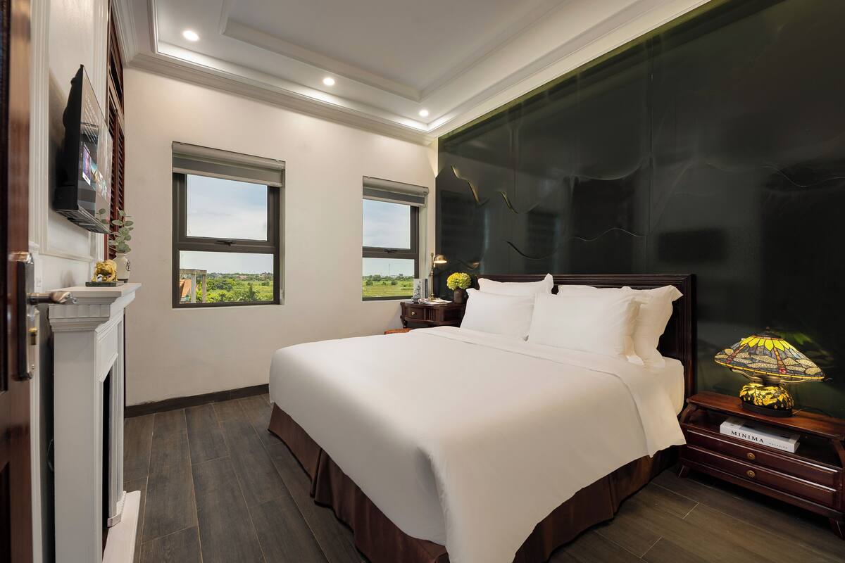 Une chambre à coucher avec un grand lit, deux fenêtres, une cheminée et une table de chevet avec une lampe.