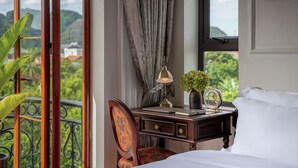 Une chambre d’hôtel avec un lit, un bureau doté d’une lampe, une chaise et une vue sur la verdure et les bâtiments, visible par la fenêtre.