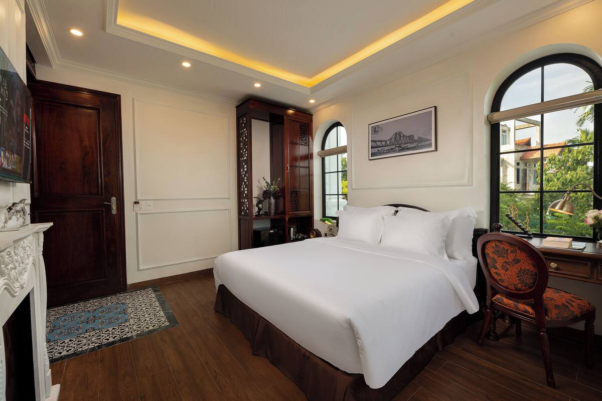 Une chambre avec un grand lit, un bureau, une chaise et une fenêtre donnant sur des immeubles et de la verdure.