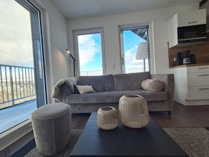 A modern living room with a grey sofa, two windows, a coffee machine, and a view of the city. - Modern vacation apartment with exceptional views and its own infrared cabin (Winterberg)