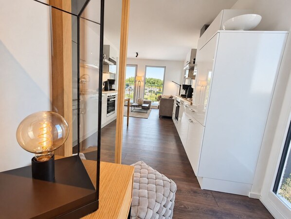 Une cuisine moderne avec des meubles blancs, un coin repas doté d’une table en bois et d’un pouf capitonné, et une vue sur l’extérieur grâce à de grandes fenêtres.