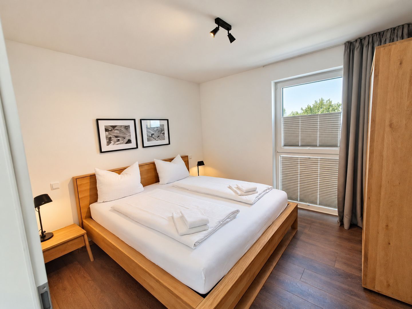 A modern bedroom with a wooden bed, white linens, two framed pictures on the wall, a bedside lamp, and a window with blinds.