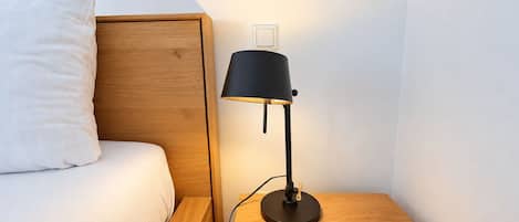 A bed with a wooden headboard, a bedside table with a lamp, and a wall-mounted light switch.