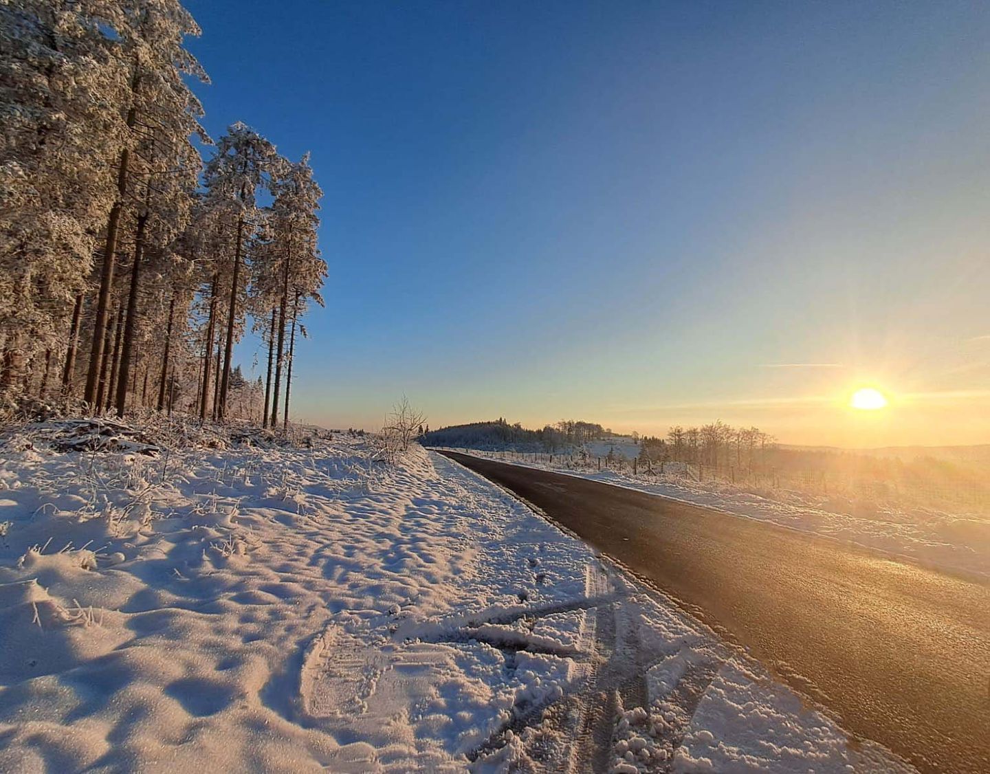Een met sneeuw bedekte weg, heldere lucht en de zon die in de verte ondergaat.