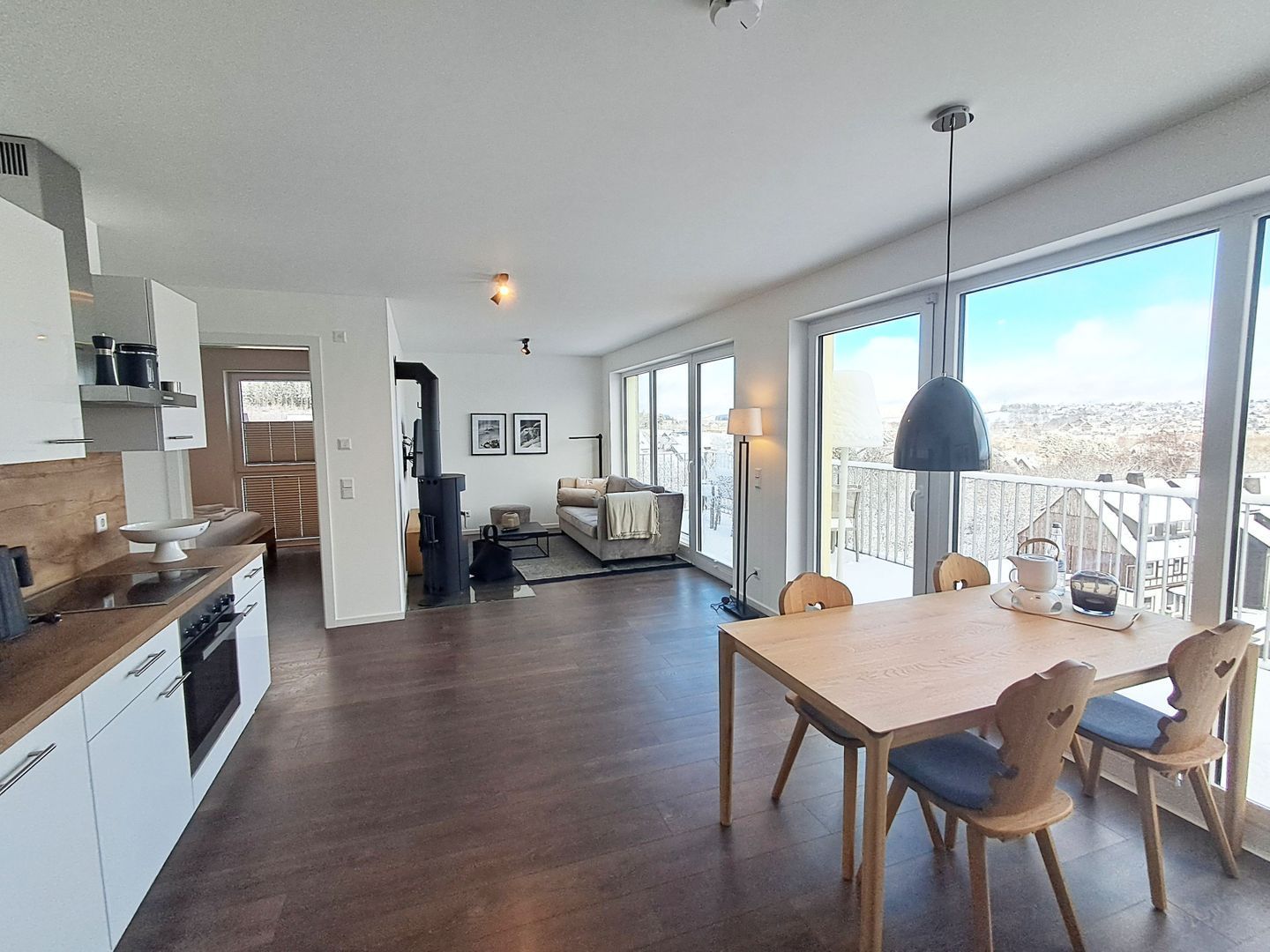 Une cuisine moderne avec un plancher en bois foncé, un coin repas avec une table et des chaises en bois, et de grandes fenêtres offrant une vue sur l’extérieur.