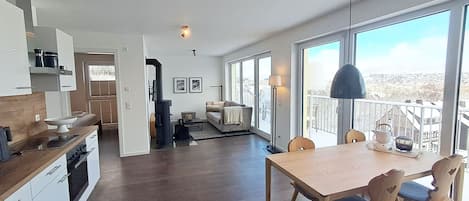 Une cuisine moderne avec un plancher en bois foncé, un coin repas avec une table et des chaises en bois, et de grandes fenêtres offrant une vue sur l’extérieur.