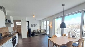 Uma cozinha moderna com soalho em madeira escura, zona de refeições com mesa e cadeiras de madeira, e grandes janelas com vista para o exterior.