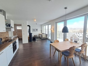 A modern kitchen with dark wood flooring, a dining area with a wooden table and chairs, and large windows offering a view of the outdoors. - Modern vacation apartment with balcony, sauna and mountain view in the mountain resort - (Winterberg)