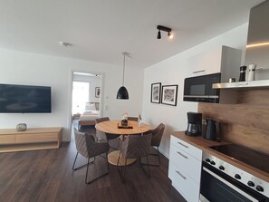 A modern kitchen with a central island, a dining area with four chairs, a flat-screen TV mounted on the wall, and a built-in oven. - Modern lodge with exceptional countryside views and private sauna - (Winterberg)