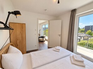 A hotel room with a bed, a desk, a chair, a lamp, and a view of the outside.