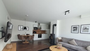 Una sala moderna con un sofá gris, una mesa de centro de madera y un comedor con una mesa redonda y sillas. Se ve una cocina con gabinetes blancos y una estufa negra al fondo.
