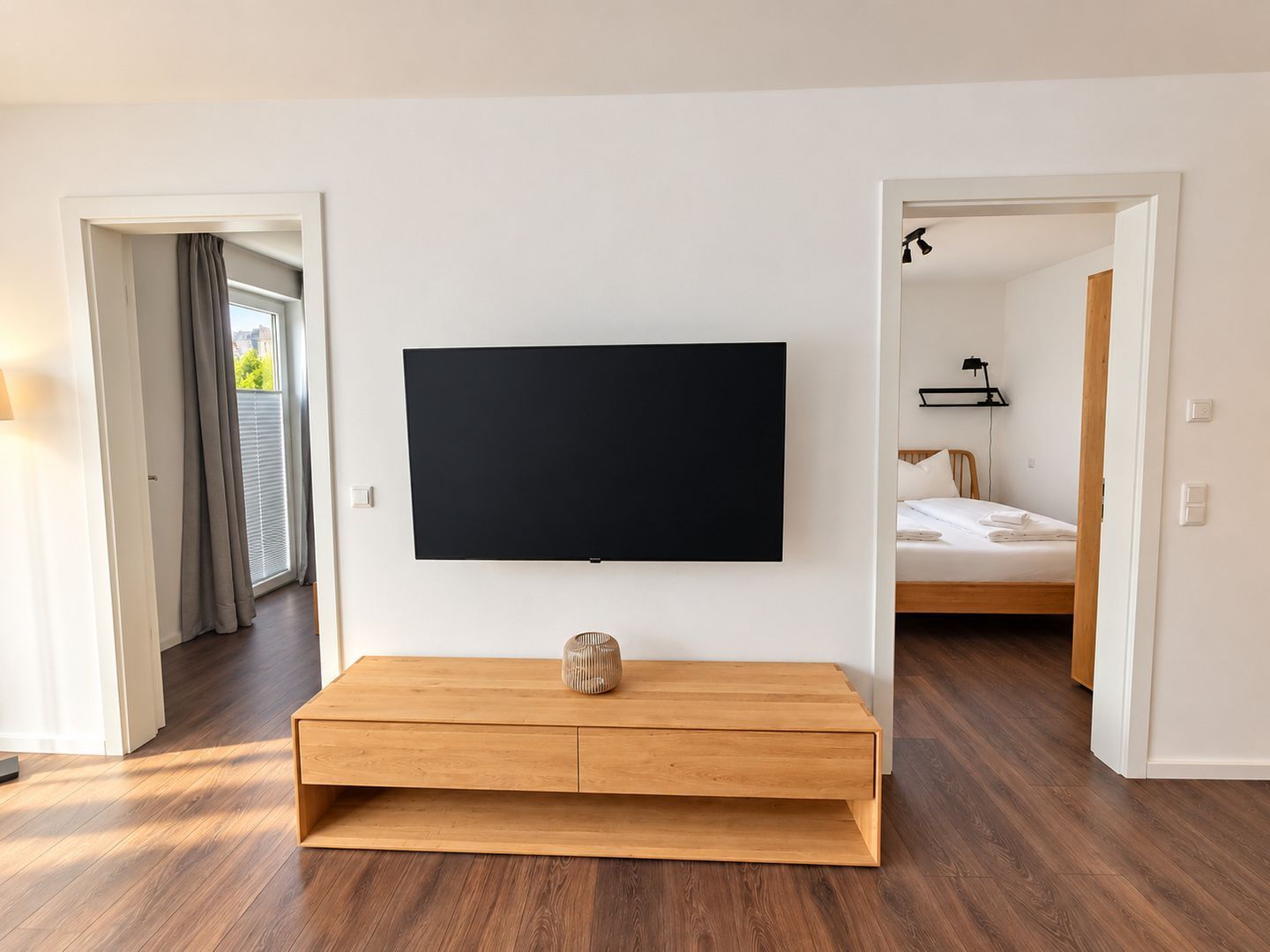 Une chambre d’hôtel moderne dotée d’un téléviseur à écran plat fixé sur une console en bois, d’un lit avec du linge de maison blanc et d’une vue sur l’extérieur par une fenêtre agrémentée de rideaux.