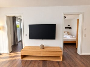 Une chambre d’hôtel moderne dotée d’un téléviseur à écran plat fixé sur une console en bois, d’un lit avec du linge de maison blanc et d’une vue sur l’extérieur par une fenêtre agrémentée de rideaux.