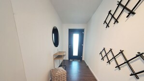 Un pasillo con suelo de madera, un reposapiés color beige, una mesita y un espejo en la pared izquierda. Se observan estanterías de metal negro en la pared derecha.