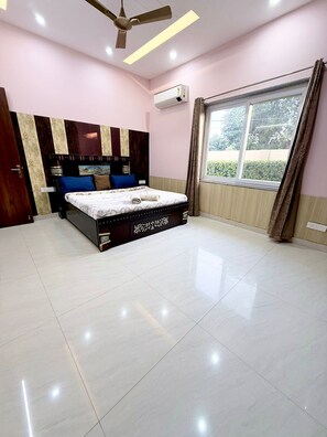 A bedroom with a bed, a ceiling fan, an air conditioning unit, a window with curtains, and a view of greenery outside.