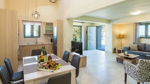A spacious living area with a dining table, chairs, a sofa, and a TV. There is a kitchen area with cabinets and a window. - Villa Iokasti with heated pool (Rethymno)