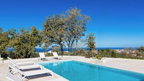 A swimming pool with lounge chairs, surrounded by trees and overlooking a coastal view. - Villa Iokasti with heated pool (Rethymno)