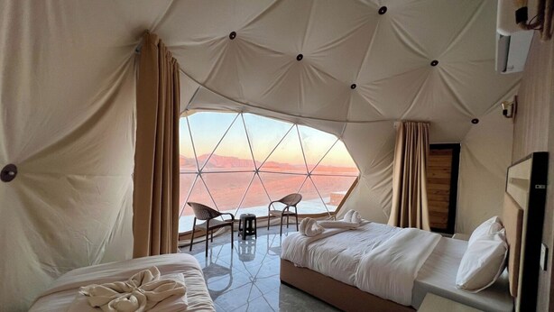 Ein Zimmer mit zwei Betten, einem kleinen Tisch und einem Stuhl, mit einem großen Fenster mit Blick auf eine Wüstenlandschaft.