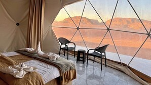 Ein geräumiges Zelt mit einem Bett, zwei Stühlen und Blick auf eine Wüstenlandschaft.