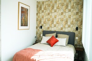 Ein Schlafzimmer mit einem Bett, Kissen, einer Decke, einer Wandleuchte und einem gerahmten Bild an der Wand.