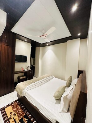 A modern bedroom with a large bed, a ceiling fan, a dark wooden wardrobe, a bedside table, and a mirror. - Kashi Darshan Hotel (Varanasi)