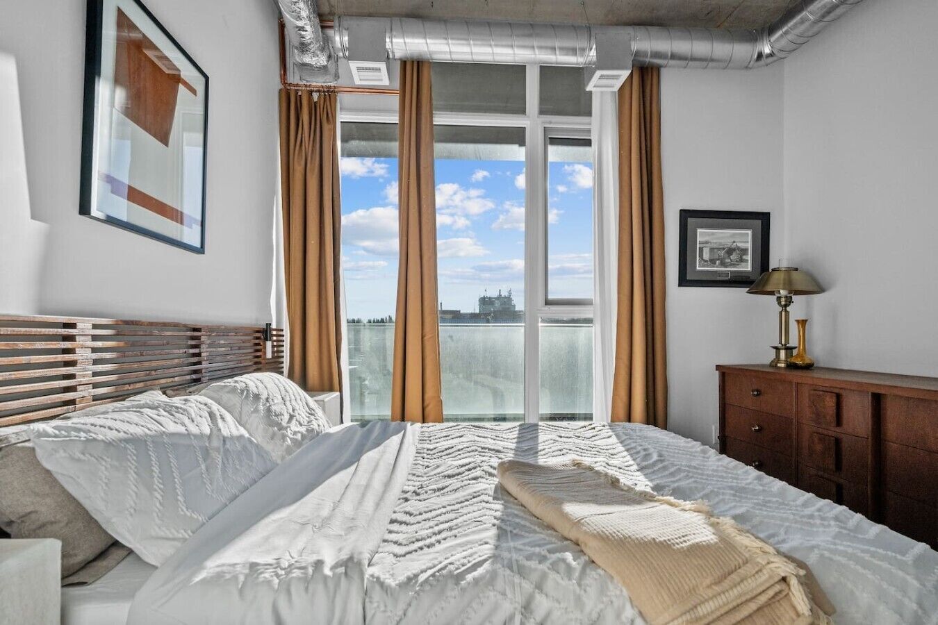 Quarto com uma cama grande, cabeceira de madeira, mesa de cabeceira, abajur e vista da cidade através de amplas janelas.