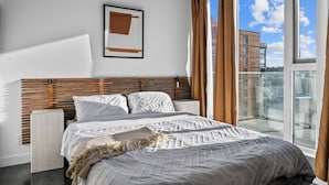 Un dormitorio moderno con una cama grande, un cabecero con listones horizontales, un cuadro enmarcado en la pared y un balcón con vistas a edificios.