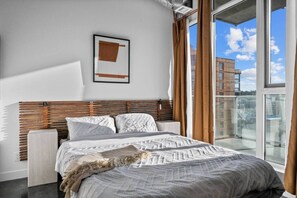Ein modernes Schlafzimmer mit einem großen Bett, einem horizontal lamellenartigen Kopfteil, einem gerahmten Bild an der Wand und einem Balkon mit Blick auf Gebäude.
