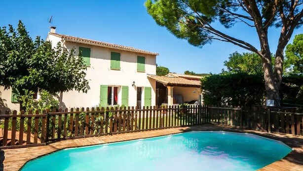 Un área de piscina con cubierta de madera, una casa con persianas verdes y un cielo azul despejado.