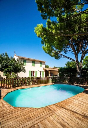 Un área de piscina con cubierta de madera, una casa con persianas verdes y un cielo azul despejado.