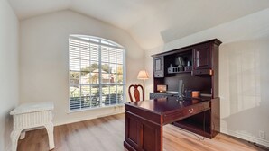 Una habitación con un escritorio de madera, una silla, un taburete blanco pequeño y un ventanal con persianas.