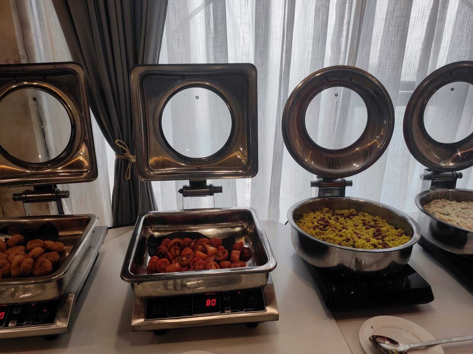 Estaciones de buffet con diversas opciones de comida, incluyendo una bandeja de salchichas, un tazón de arroz y una olla que parece ser un pastel de carne.