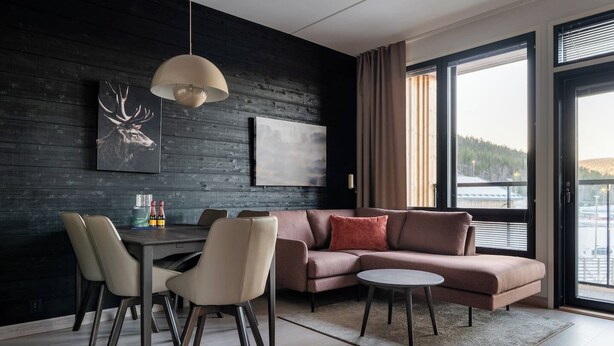 A modern living room with a dark wooden wall, a sectional sofa, a dining table with chairs, and a large window offering a view of the outdoors. - Le Chalet Lingonberry D6 by Aavalevi (Levi)