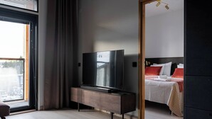 A modern hotel room with a large flat-screen TV on a wooden cabinet, a bed with red and white bedding, and a view of the city through a large window. - Le Chalet Lingonberry D6 by Aavalevi (Levi)