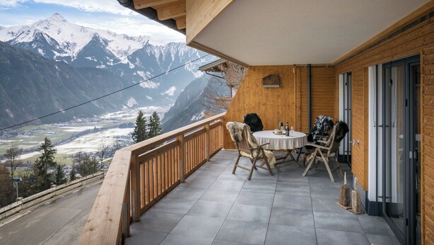 A wooden balcony with a table and chairs overlooking a snowy mountain range.