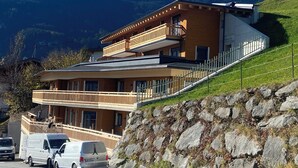 A modern two-story building with a balcony, situated on a hillside with a mountain backdrop.