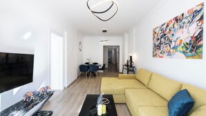 Een moderne woonkamer met een grote flatscreen televisie, een gele bank, een salontafel en een eethoek met stoelen en een tafel.