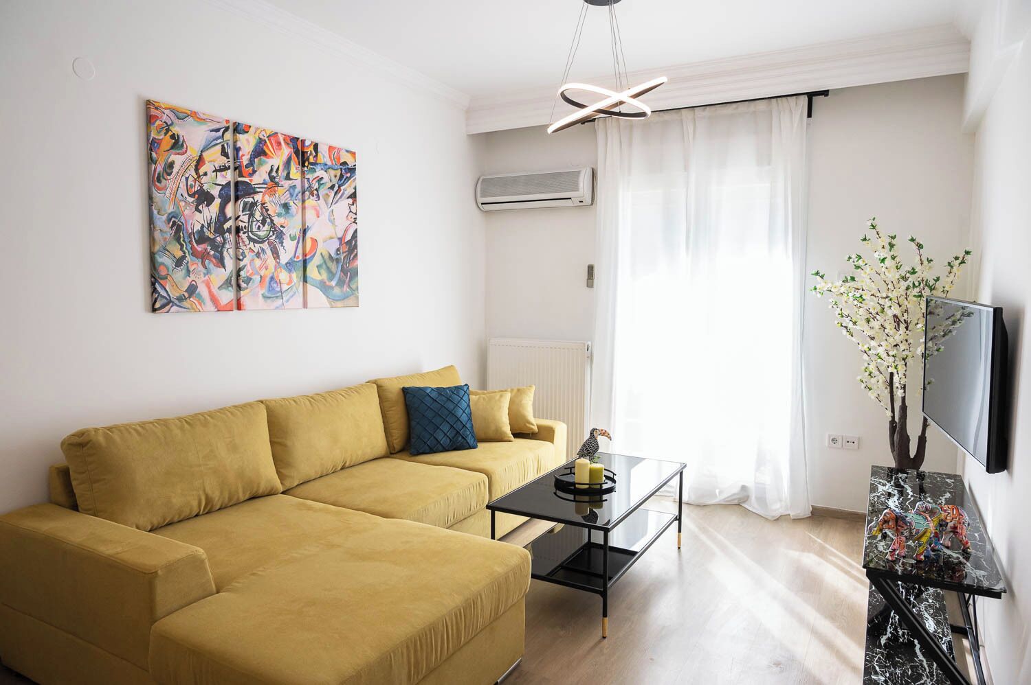 A living room with a mustard sofa, a glass coffee table, a colorful abstract painting, a modern light fixture, and a TV on a black stand.