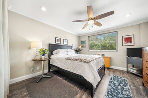 Een slaapkamer met een bed, een plafondventilator, een raam, een televisie en een dressoir.