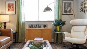 Un soggiorno con un divano in pelle, un tavolino da caffè in vetro con una ciotola verde, una poltrona bianca, una lampada, un segnale che recita “ATLANTA” e una pianta.