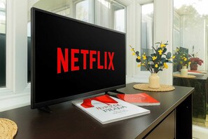 A television with the Netflix logo on a dark surface, a vase with yellow flowers, and a stack of books.