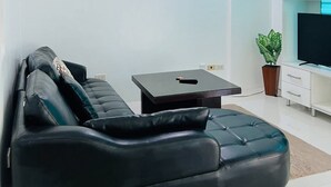 A modern living room with a black leather sofa, a small black coffee table, and a flat-screen TV on a white cabinet. - Cozy 1 bedroom condo inside Subic Bay Freeport Zone with complete amenities. (Subic Bay Freeport Zone)