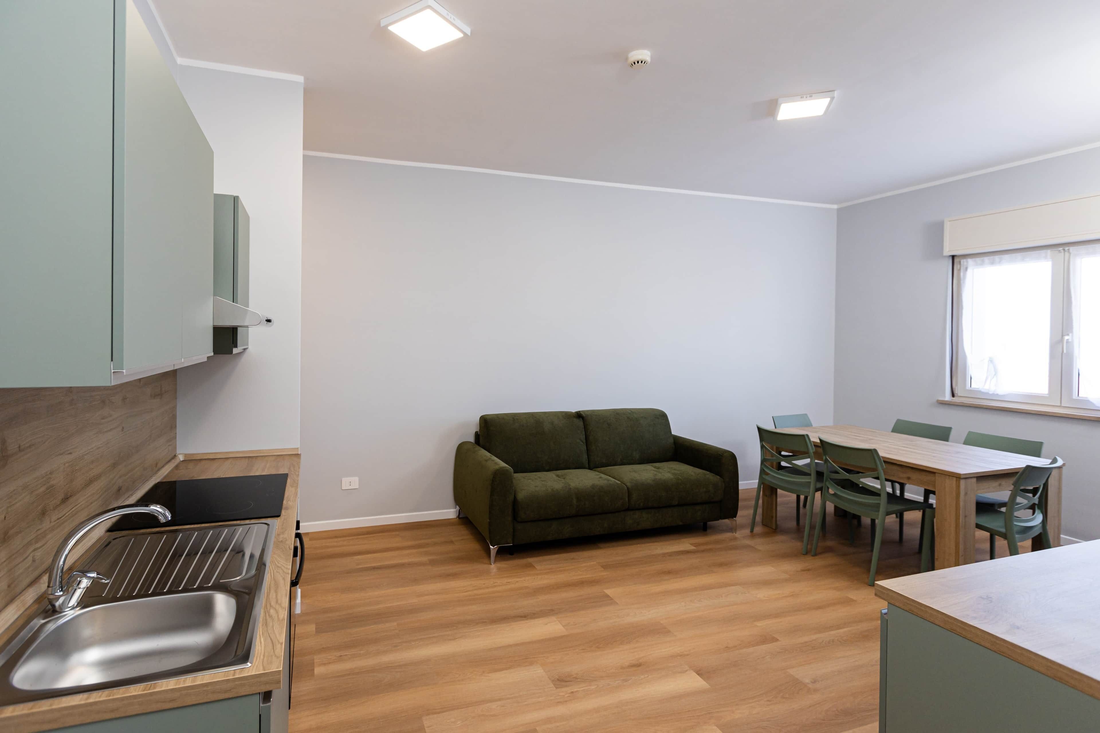 A compact kitchen with a sink, a green sofa, a dining table with chairs, and a window with curtains.
