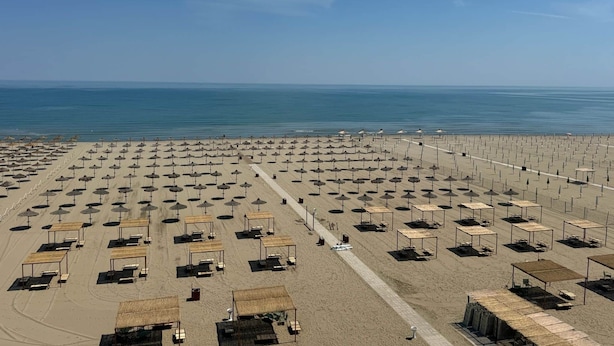 Ein Strand mit Reihen von Sonnenschirmen und Liegestühlen, einem klaren Himmel und dem Meer im Hintergrund.