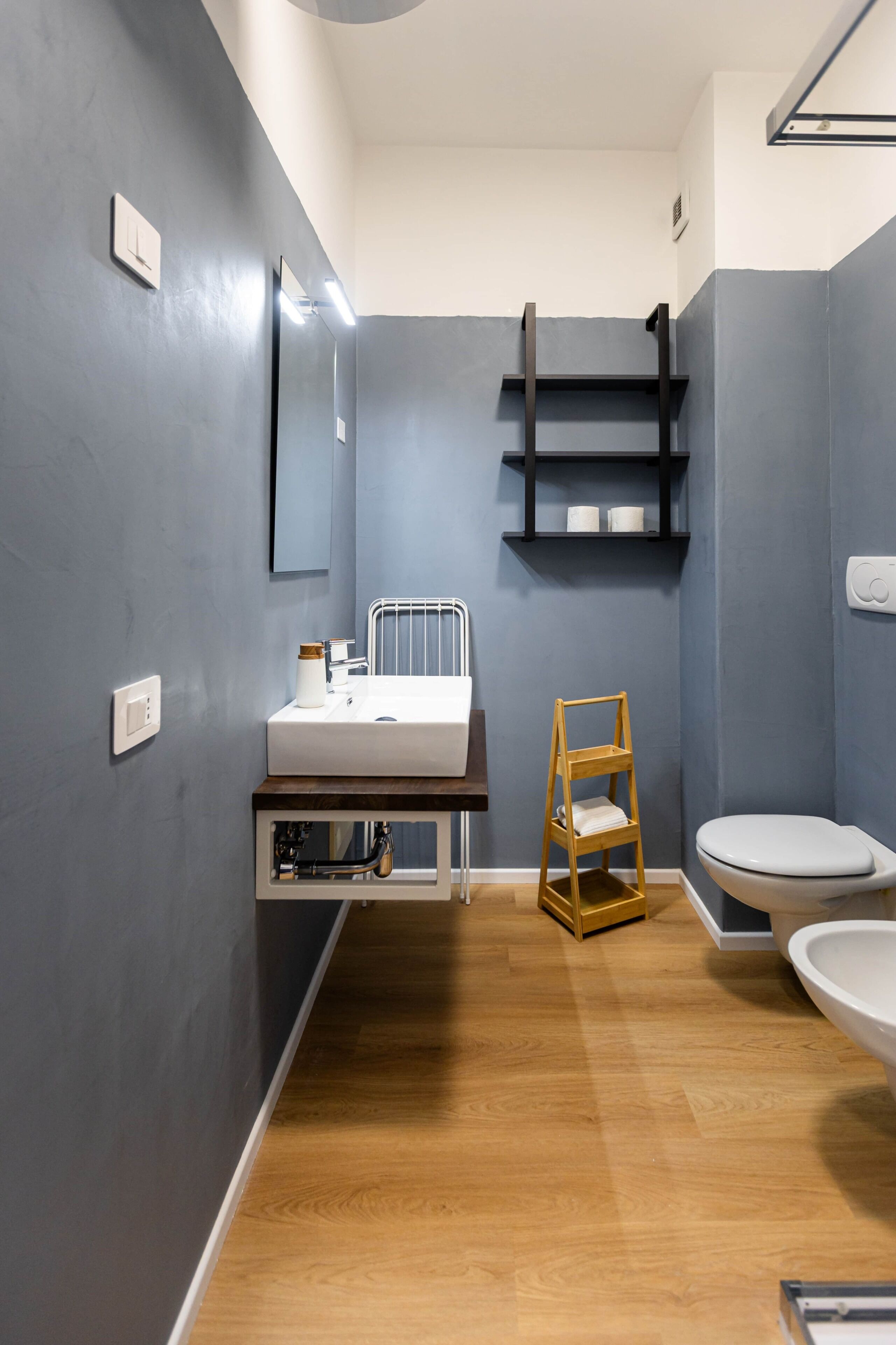 Une salle de bain avec un plancher en bois, un lavabo blanc, des toilettes, un miroir et une étagère contenant des articles de toilette.