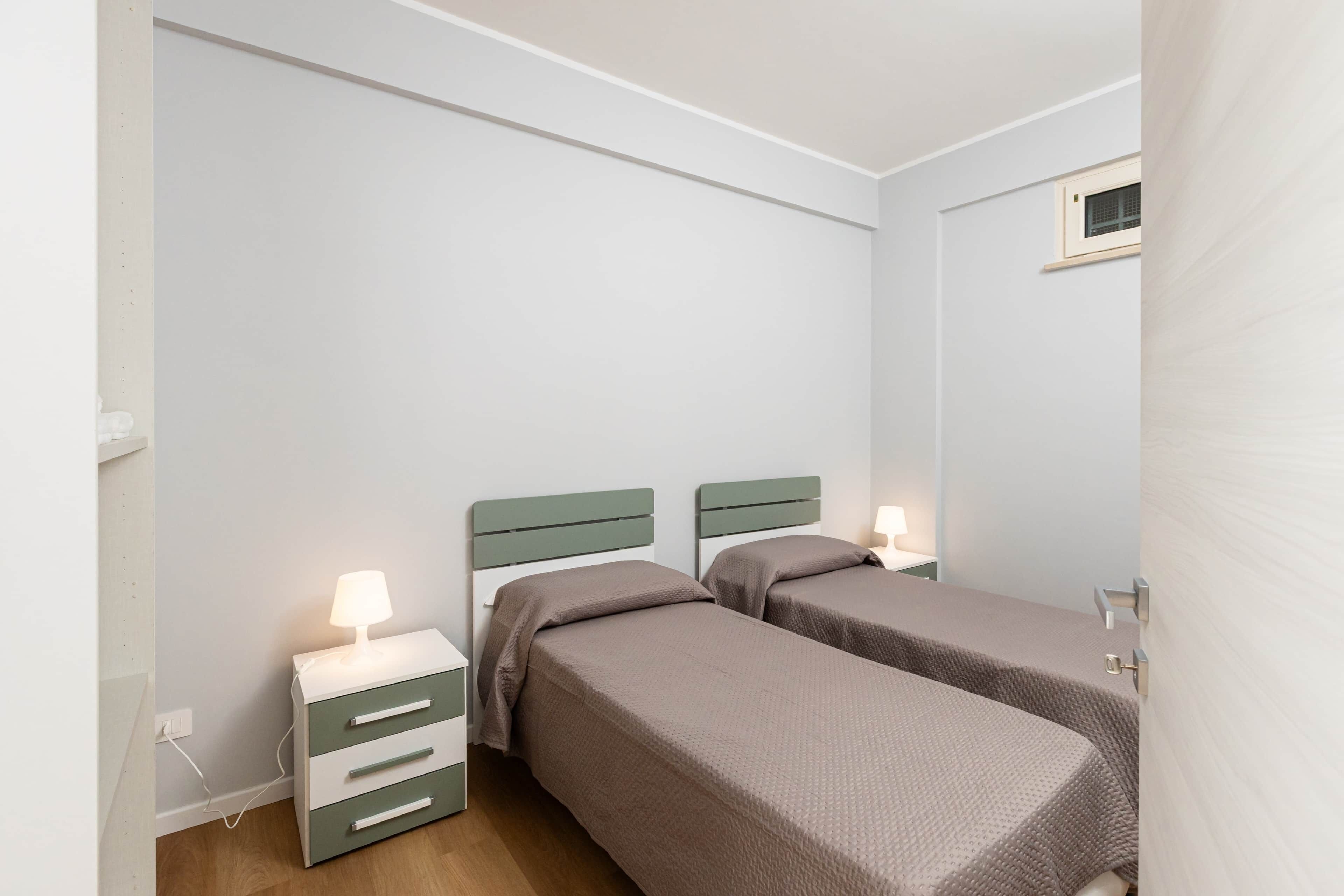 A small, clean room with two single beds, a bedside table with a lamp, and a power outlet.