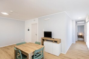 Une pièce moderne et bien éclairée, avec une table et des chaises de salle à manger en bois, un petit téléviseur posé sur un meuble en bois et un buffet blanc.