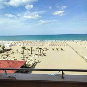 Ein Strand mit Reihen von Sonnenschirmen und Liegestühlen, einem klaren Himmel und dem Meer im Hintergrund.