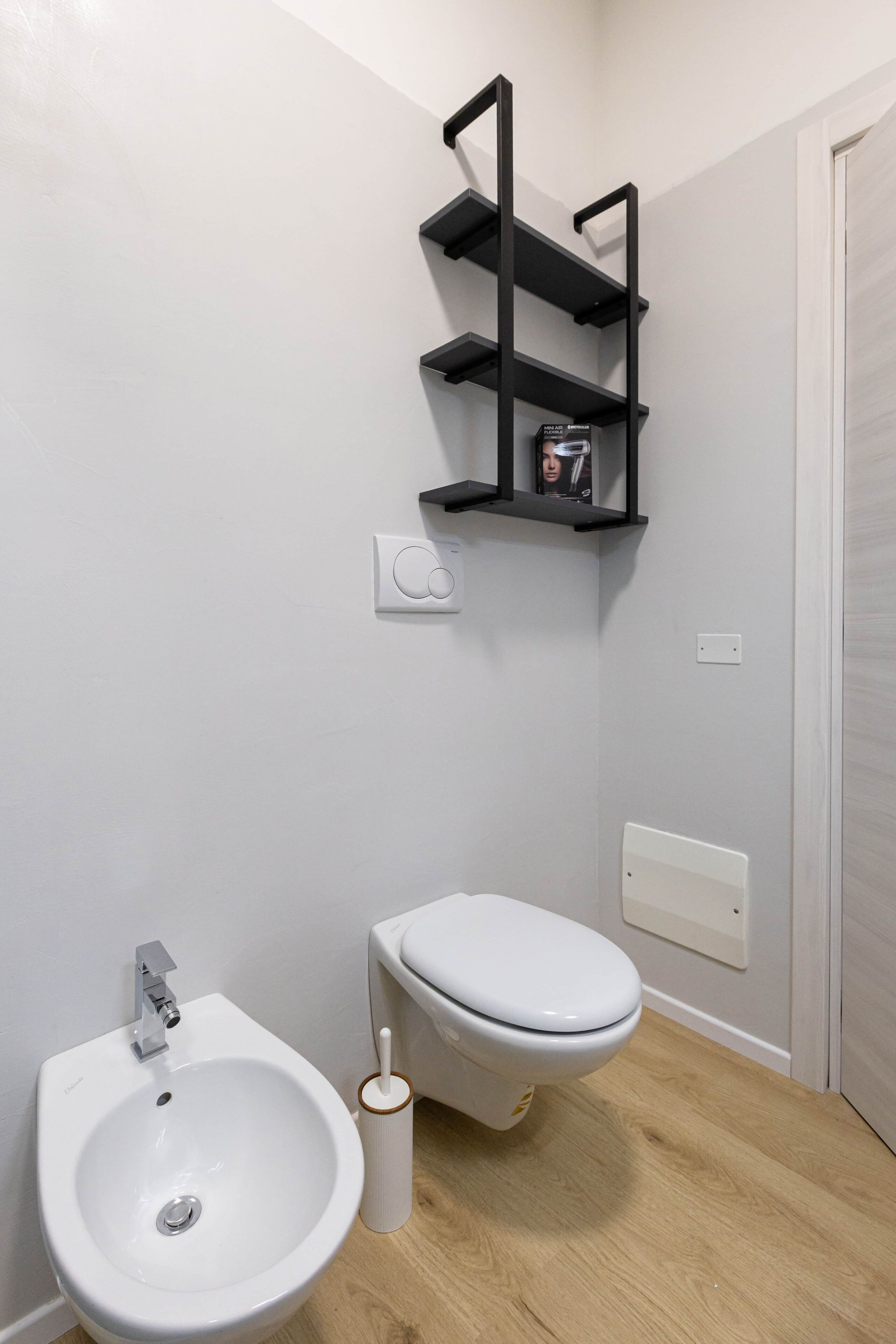 A bathroom with a white bidet, a white toilet, and a black shelf unit above the toilet.